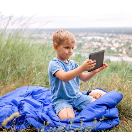 Local vacation. Cute boy having rest in sleeping bag on the top of the mountain not far from town and chatting with friends via internet, outdoor summer weekend, social distance lifestyleの写真素材