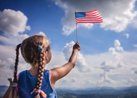 Patriot and National Flag day celebration. Cute little patriot sitting on the meadow and holding the national flag of United States over blue sky background, summer outdoor, soft focus on flagの写真素材