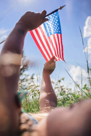 Patriot and National Flag day celebration. Cute little patriot sitting on the meadow and holding the national flag of United States over blue sky background, summer outdoor, soft focus on flagの写真素材