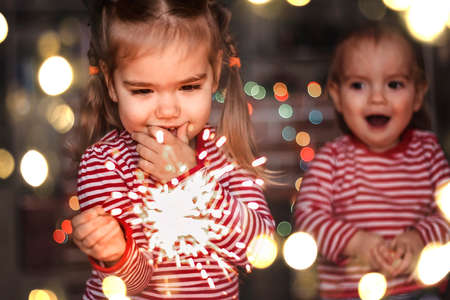 Family Christmas at home. Two happy kids, toddler boy and cute small girl in Santa hat holding burning sparkler and smile happily over defocused Christmas background, indoor emotional lifestyleの写真素材