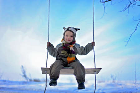 Cute toddler boy in funny hat and warm coat staying alone on the frozen lake at winter walk outdoors, winter family lifestyleの写真素材