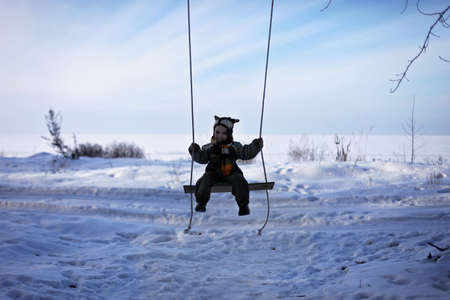 Cute toddler boy in funny hat and warm coat staying alone on the frozen lake at winter walk...