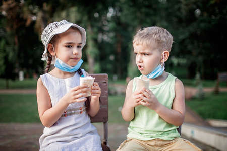 Cute sibling with face mask wearing down eating ice cream during quarantine, outdoor closeup portrait, hot weather in new normal realityの写真素材