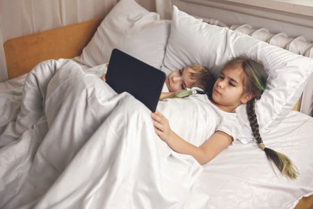 Two siblings watching video in their mobile electronic device in the bed, gadget dependence and addiction, online and internet for the whole day, family lifestyle, indoorの写真素材