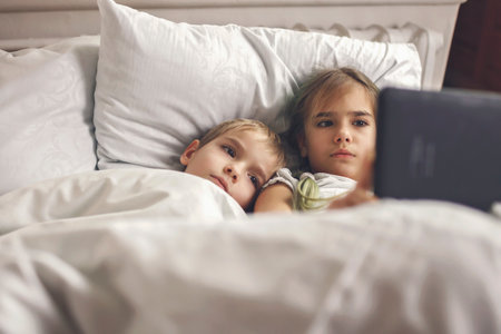 Two siblings watching video in their mobile electronic device in the bed, gadget dependence and addiction, online and internet for the whole day, family lifestyle, indoorの写真素材