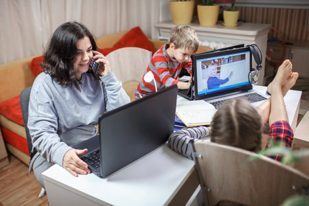 Distant education and work at home, children doing homework, mother working and help them. Elementary school kids during online class with parent working remotely in one room, lockdown, focus on womanの写真素材
