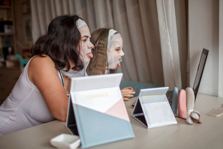 Beautiful girl and her mother using gadgets during spa procedure, mom and daughter chatting via internet with smooth mask on face, wellness, relaxation and beauty salon at home, indoor lifestyleの写真素材