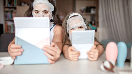 Beautiful girl and her mother watching beauty master class with tablets online and doing spa procedure by themselves, mom and daughter having fun with smooth mask on face, beauty salon at homeの写真素材