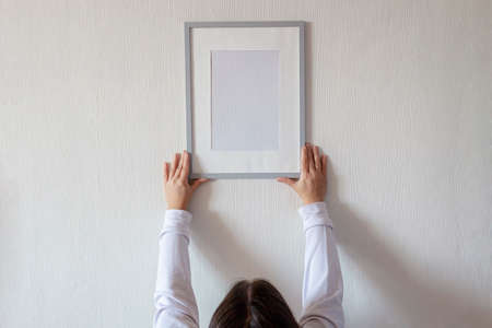 Woman in white hoody hanging vertical empty frame on white wall to create family photo gallery, to...