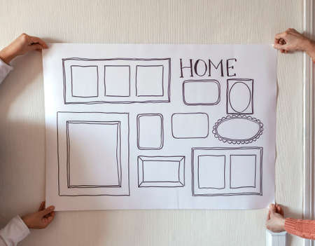Woman evaluating and sizing up white poster with drawn set of different empty vertical and horizontal picture frames on white wall, planning family photo gallery project to capture moment, lifestyleの写真素材