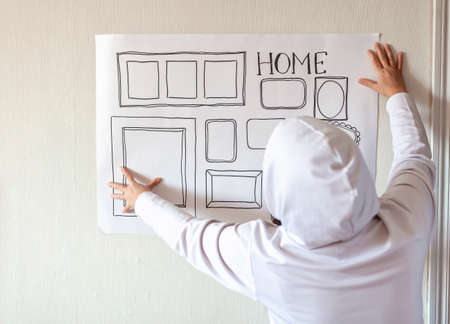 Woman evaluating and sizing up white poster with drawn set of different empty vertical and horizontal picture frames on white wall, planning family photo gallery project to capture moment, lifestyleの写真素材