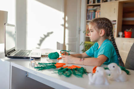 Elementary school girl doing carrot with wooden clothes pins for Easter celebration looking video on laptop, online workshop, kid at-home activity, creativity and distant education, holiday decorationの写真素材