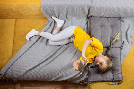 Digital native generation alpha, cute kid lying on yellow sofa with gray blanket and using smartphone for chatting and distant learning, online education at home, new normal lifestyle, trendy colorsの写真素材