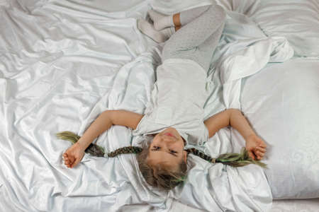 Cute awaking girl in white clothes with two braids lying on the bed in bedroom in the morning, happy family concept, healthy lifestyle and sleep careの写真素材