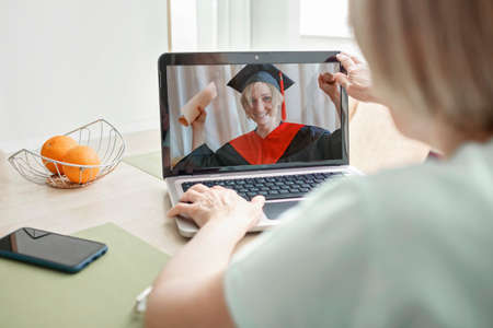 Virtual graduation and convocation ceremony. Senior woman congratulating her daughter in graduation gown and cap during online video call, distant education andの写真素材
