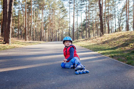 Sad kid in sport helmet sitting on road during riding on roller skates at park, active family...