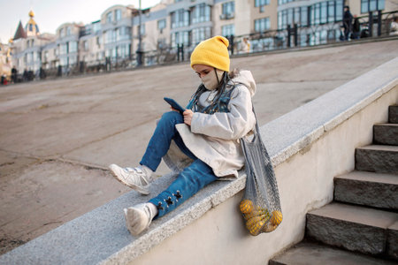Preteen girl in medicine mask texting with smartphone on street stairs on way from shopping, trendy schoolgirl with grey mesh eco bag with purchase, yellow fruits, zero waste, sustainable lifestyleの写真素材