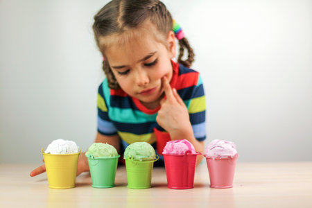 Pretty little girl eating an ice-cream from some different colorful buckets over white background, food and drink concept, portrait studio shotの写真素材