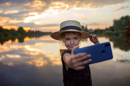 Pretty preteen girl in summer straw hat taking selfie for cellphone at beautiful sunset on lake, lifestyle, local travel concept, young bloggerの写真素材