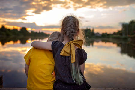 Pretty preteen girl with her younger brother looking at beautiful sunset on lake, lifestyle, happy childhood, family travel, back viewの写真素材