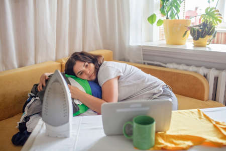 Tired middle aged woman watches video on tablet and ironing things at the same time, daily routine, social media addiction, housewife and mom burnout conceptの写真素材