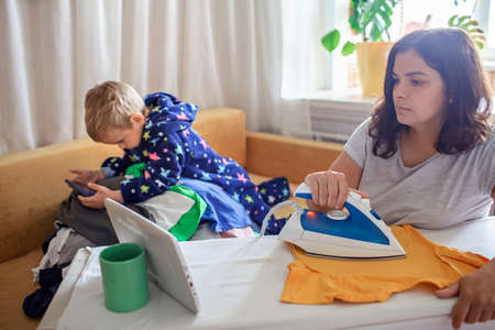 Tired middle aged mom watches video on tablet and ironing things at the same time, next to her son with smartphone too, social media and gadget addiction, housewife and mom burnout conceptの写真素材