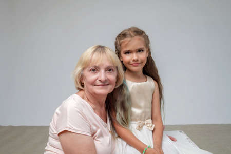 Happy preteen girl with long hair in festive dress with her senior granny over white pastel...