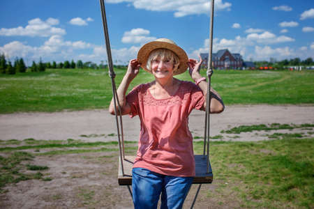 Carefree senior woman in straw hat swinging on a wooden swing and looking at the camera, mental and physical health, age beauty, baby boomer generation, summer lifestyleの写真素材