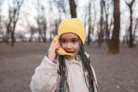 Fashionable pretty girl in yellow hat and grey coat talking on a banana as if a cell phone in the autumn forest, beauty and fashion, healthy eating, fun and digital detox joke, outdoors lifestyleの写真素材