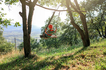 Summertime after lockdown. Cute little boy sitting alone on a handmade swing in the mountains far from usual playground, outdoor summer lifestyleの写真素材
