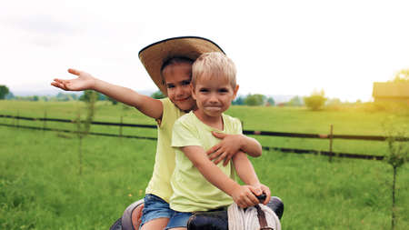 Little girl in cowboy hat and her smaller brother playing in western in the farm among mountains, original vintage saddle, happy summertime in the countryside, childhood and dreamsの写真素材