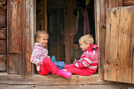 Happy boy and girl having fun and rest together on the doorstep of old wooden house during summer vacation, eco-friendly travel concept, outdoor summertime lifestyleの写真素材