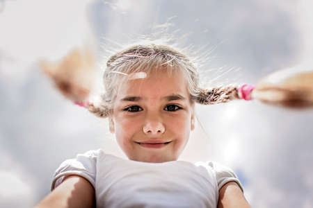 Close-up candid portrait of cute 7-8 years old girl with two funny braids taken up at the sky from the ground, happy summertime, outdoor lifestyleの写真素材