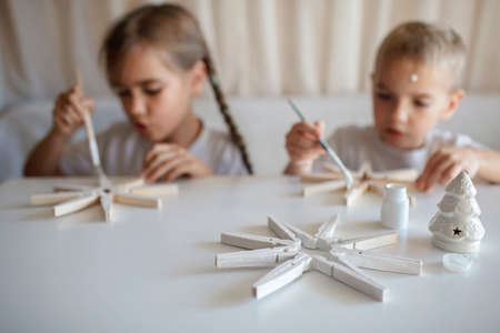 Siblings, boy and girl, paint wooden snowflakes from clothespins with white paint at home, hands in paint, recyclable craft ornament, New Year DIY decoration, selective focus on snowflakeの写真素材