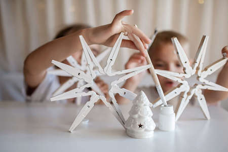 Siblings, boy and girl, paint wooden snowflakes from clothespins with white paint at home, hands in paint, recyclable craft ornament, New Year DIY decoration, selective focus on snowflakeの写真素材