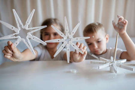 Siblings, boy and girl, paint wooden snowflakes from clothespins with white paint at home, hands in paint, recyclable craft ornament, New Year DIY decoration, selective focus on snowflakeの写真素材