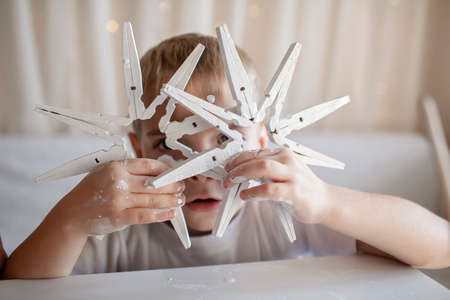Little boy in white t-shirt paints wooden snowflakes from clothespins with white paint at home, hands in paint, recyclable ornament, New Year DIY decoration, monochrome photography, focus on craftの写真素材