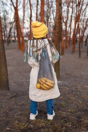 Teenager girl carries gray eco mesh bag with bright yellow fruits through the autumn park after...