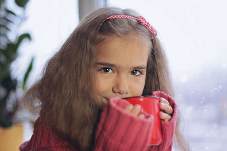 Child in warm comfort sweater drinking a cup of hot chocolate, relaxing on a window sill and looking...
