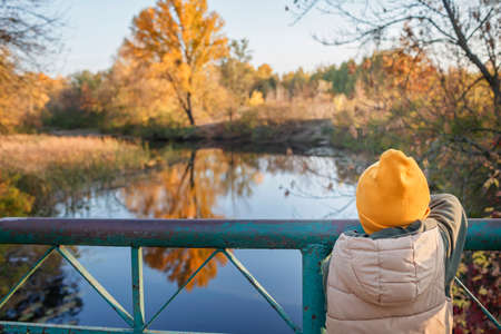 Pensive little boy stands on bridge and admires the magnificent view of autumn forest and its reflection in the lake water. Autumn walk. Outdoor lifestyle, active family lifestyle, view from behindの写真素材