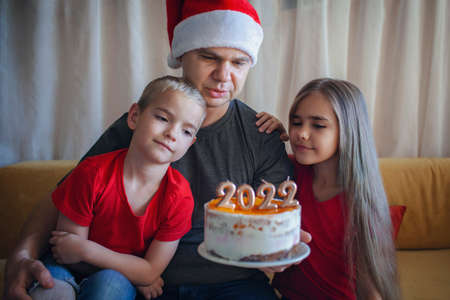 Happy dad with two children looks at the candles on Christmas cake with golden 2022 numbers on top with yellow icing over bokeh lights background, family New Year celebration, focus on fatherの写真素材
