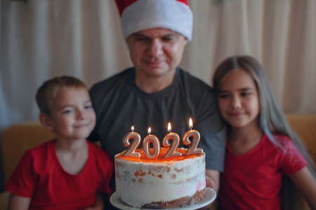 Dad with two children looks dreamily at burning candles on Christmas cake with golden 2022 numbers on top with yellow icing over bokeh lights background, family New Year celebration, focus on candlesの写真素材
