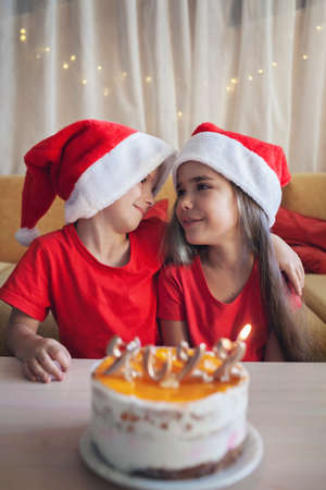 Happy siblings hug each other before Christmas cake with burning golden 2022 candles on top with yellow icing over bokeh lights background, family New Year celebration, focus on kidsの写真素材