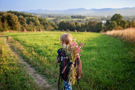 Little hiker with backpack comes from the mountain and carries a bouquet of wildflowers that he collected for his mother, gorgeous landscape, beauty of nature, view from the backの写真素材