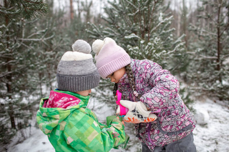 Happy kids with cup of tea covered with plaid during winter picnic in forest on snowing day, outdoor walking, excited family in warm clothesの写真素材