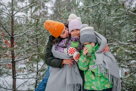 Happy kids with cup of tea covered with plaid during winter picnic in forest on snowing day, outdoor walking, excited family in warm clothesの写真素材