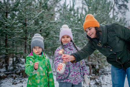 Happy kids with burning sparklers during walking in forest on snowing day, outdoor family Christmas celebration, excited family in warm clothesの写真素材