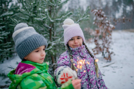 Happy kids with burning sparklers during walking in forest on snowing day, outdoor family Christmas celebration, excited family in warm clothesの写真素材