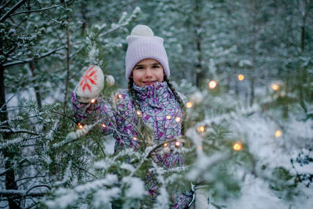 Outdoor portrait of beautiful happy girl, smiling and laughing, plays with glowing garland during walking in forest on snowing day, family weekendの写真素材
