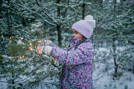 Outdoor portrait of beautiful happy girl, smiling and laughing, plays with glowing garland during walking in forest on snowing day, family weekendの写真素材